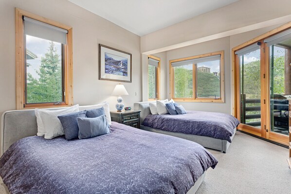 A bedroom with two beds, a nightstand, a lamp, and large windows with a view of greenery and a building.
