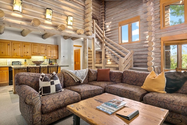 Interior de una cabaña de troncos con una escalera de madera, un sofá grande y una zona de cocina.