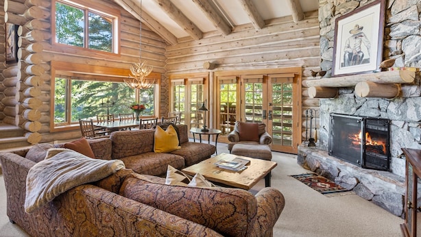 L’intérieur d’une cabane en rondins, avec une cheminée en pierre, de grandes fenêtres et un coin salon confortable.