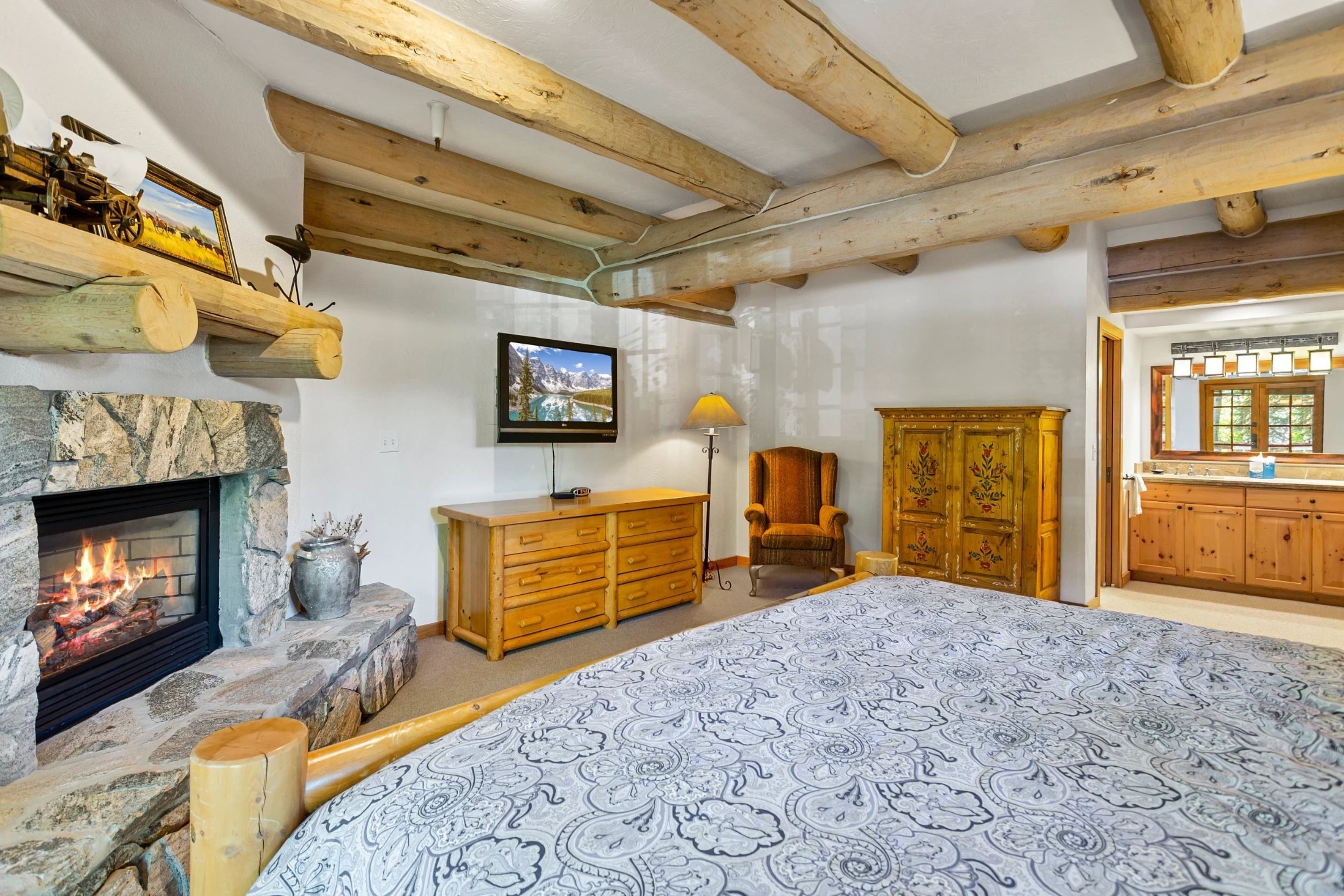Ein gemütliches Zimmer mit Kamin aus Stein, Holzbalken, einem Bett mit gemusterten Betttextilien, einem Fernseher, einem Holzschrank und einem Sessel.