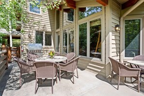 Un patio exterior con mobiliario de mimbre, una parrilla y ventanas amplias con vistas a una zona boscosa.