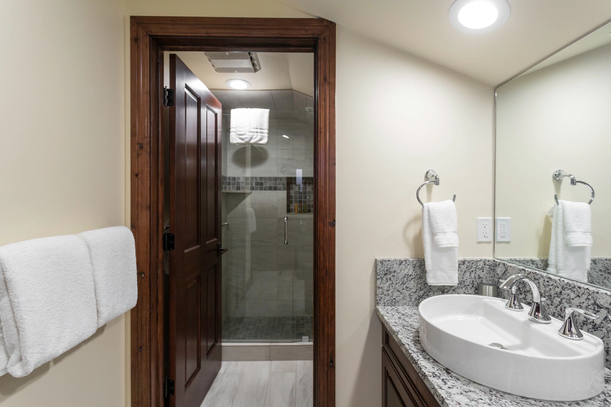 Un baño con puerta de madera, cabina de ducha de vidrio, lavamanos con dos grifos y un espejo.