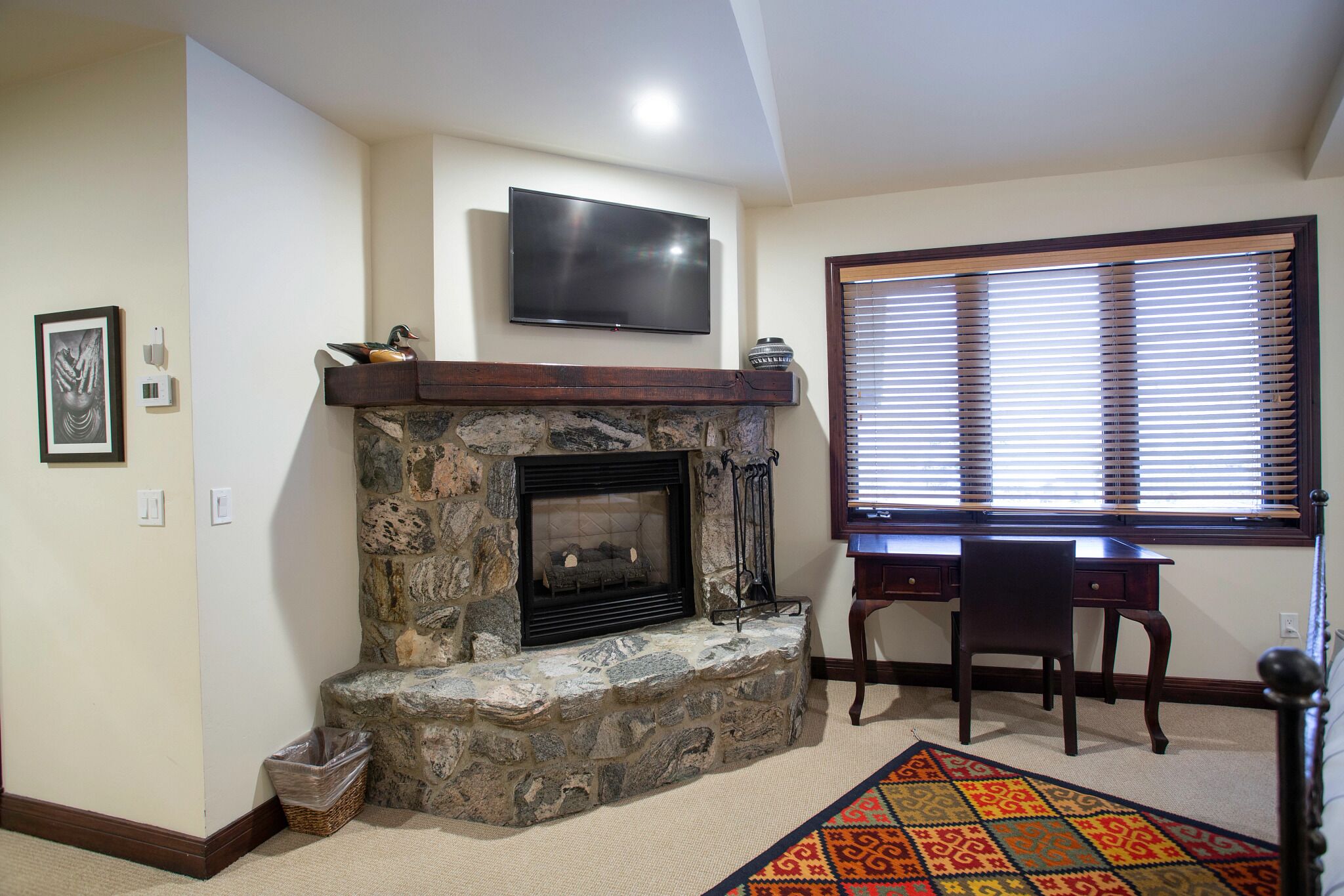 A living room with a stone fireplace, a flat-screen TV mounted above it, a desk with a chair, and a window with blinds.