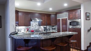 A kitchen with dark wooden cabinets, a granite countertop, a stainless steel range hood, a microwave, and a toaster oven. There are three stools at the island.