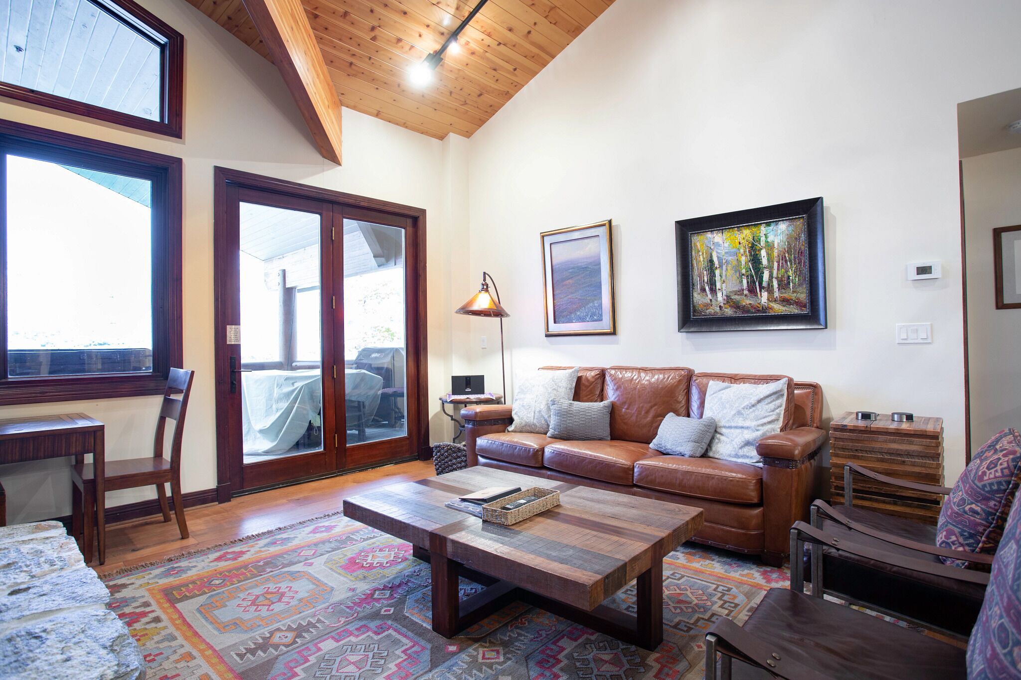 A living room with a brown leather sofa, a wooden coffee table, a patterned rug, and a large window with a view of the outdoors.