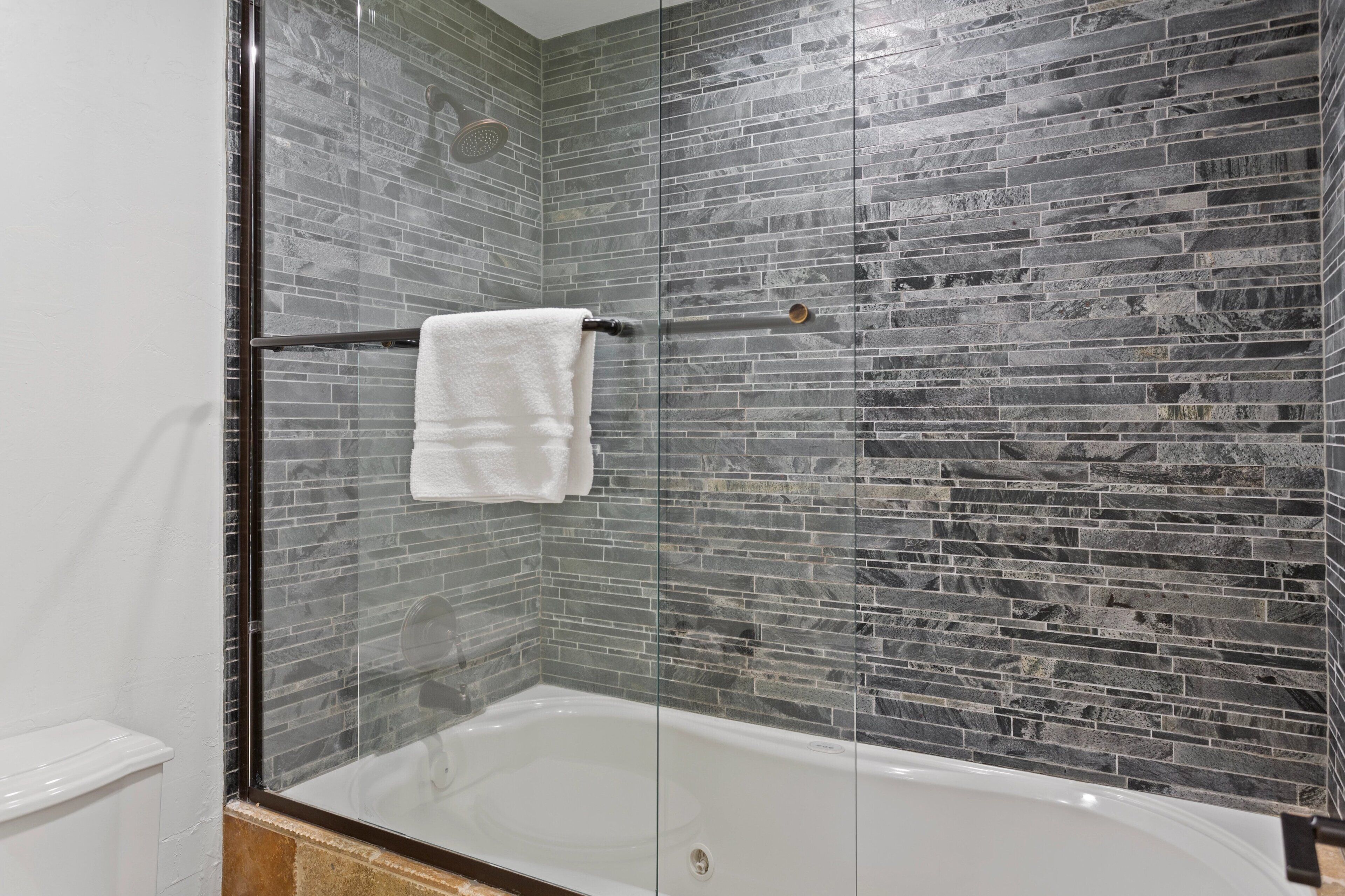 A modern bathroom with a glass-enclosed shower, a white bathtub, and a tiled wall with a stone-like pattern.