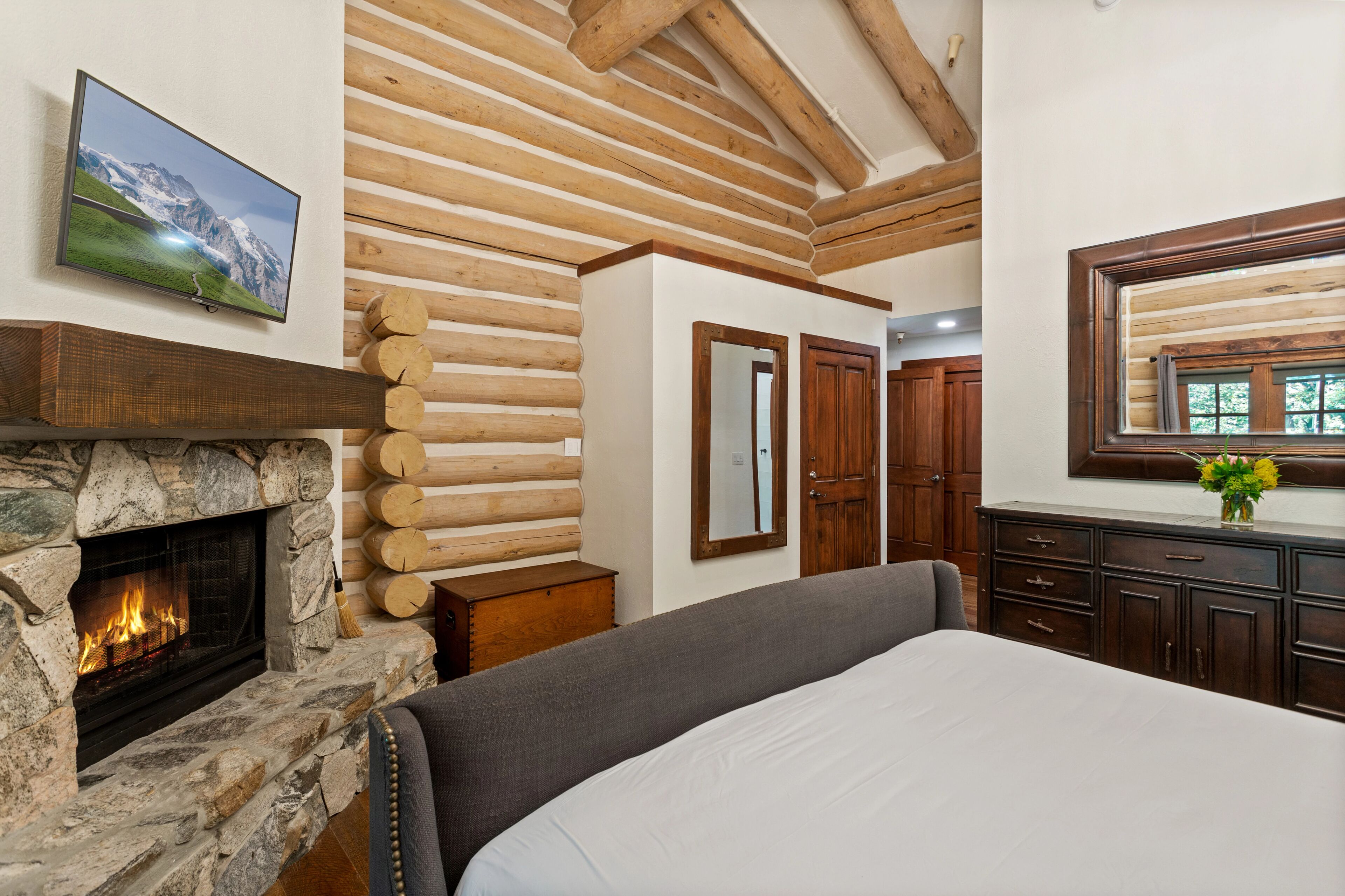Une chambre avec une cheminée en pierre, un lit, un plafond en bois et un miroir au-dessus d’une commode.