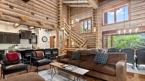 A log cabin interior with a wooden staircase, a living area with a sofa and coffee table, and a kitchen with dark cabinets.