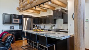 A modern kitchen with dark cabinets, stainless steel appliances, a marble island with bar stools, and wooden ceiling beams.