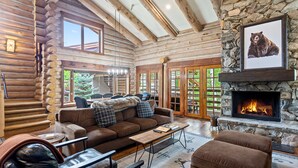 A log cabin interior with a stone fireplace, wooden furniture, and a large window overlooking greenery.