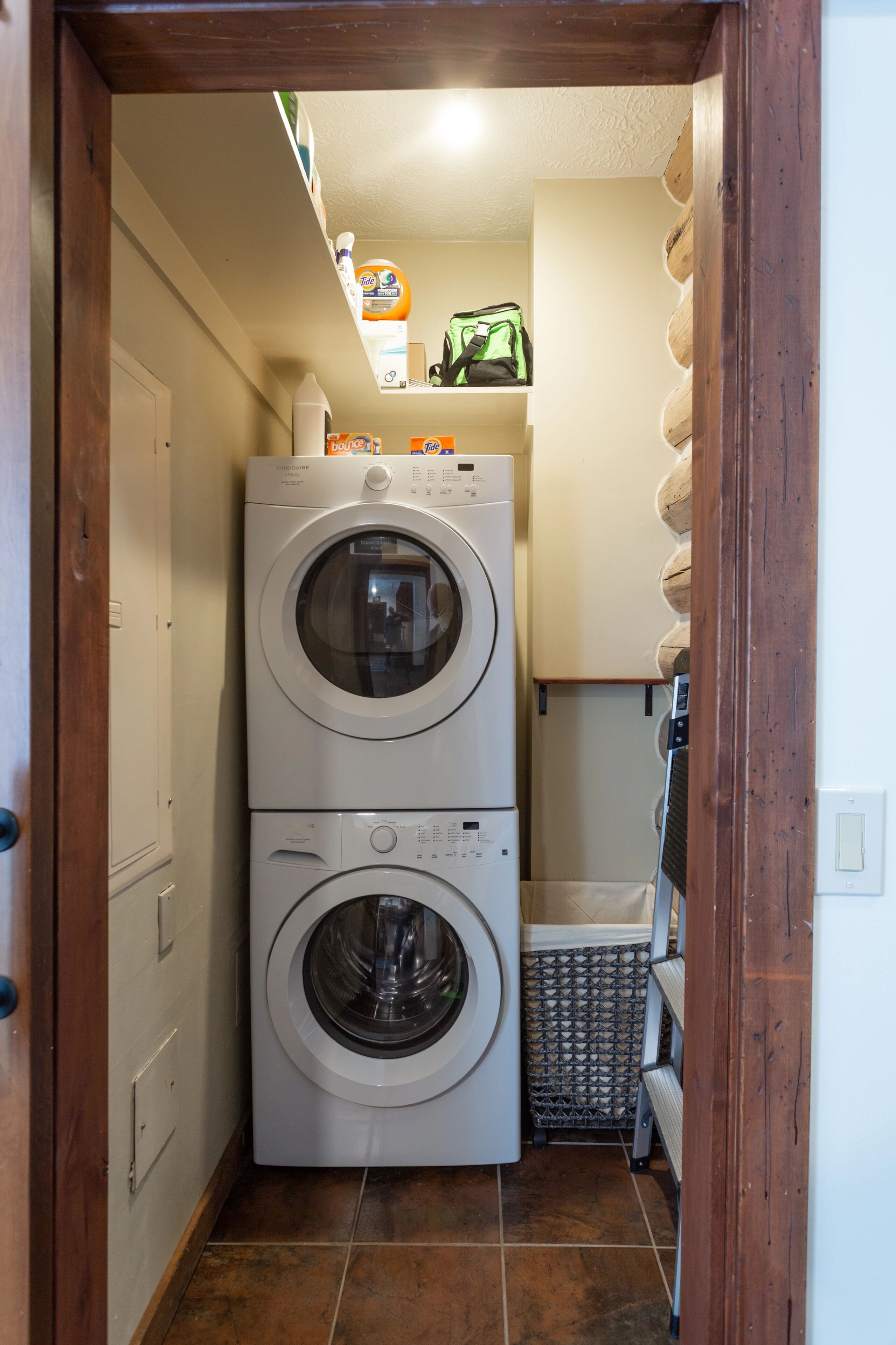 A laundry room with stacked washers and dryers, a small shelf with cleaning supplies, and a tiled floor.