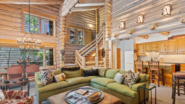 A log cabin interior with a green sectional sofa, a dining area, a kitchen with wooden cabinets, and a staircase.