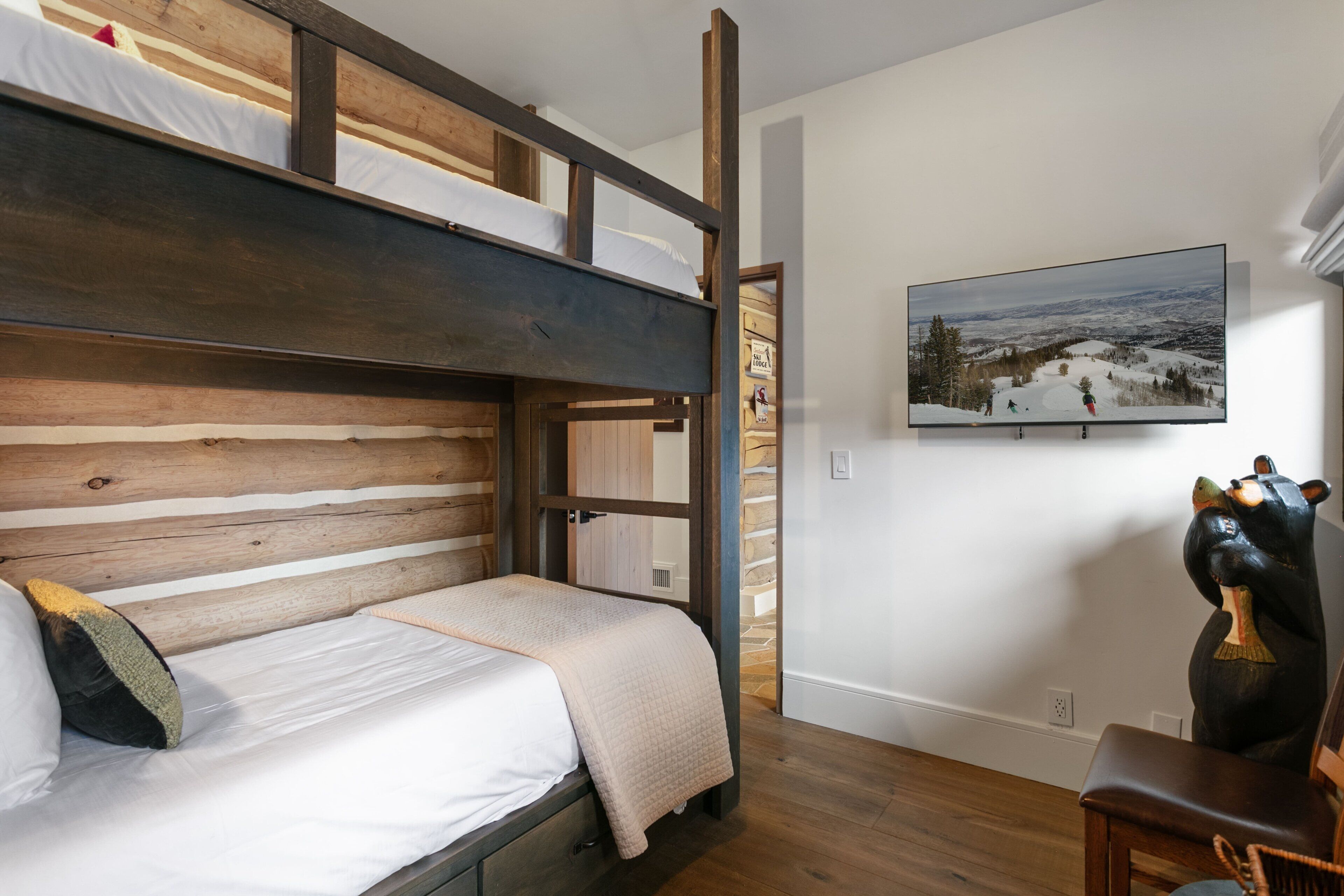 Una cama litera con cabecera de madera, un televisor montado que muestra un paisaje nevado y una escultura de oso sobre una mesa.