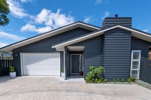 A modern two-story house with a dark siding exterior, a white garage door, and a small front porch with a potted plant. - Inn McDowell - Modern 3-Bed, Minutes to Te Puia (Rotorua)