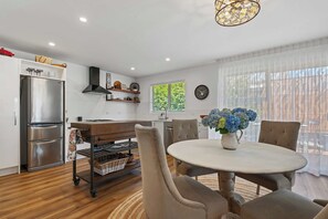 A modern kitchen with a stainless steel refrigerator, a wooden dining table with chairs, and a large window with blinds.