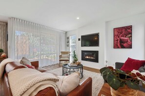 A living room with a fireplace, a sofa, a coffee table, and a large window with sheer curtains.