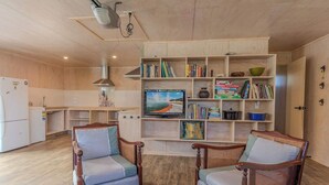 Een woonkamer met een houten boekenkast, twee fauteuils, een televisie en een keukenhoek.