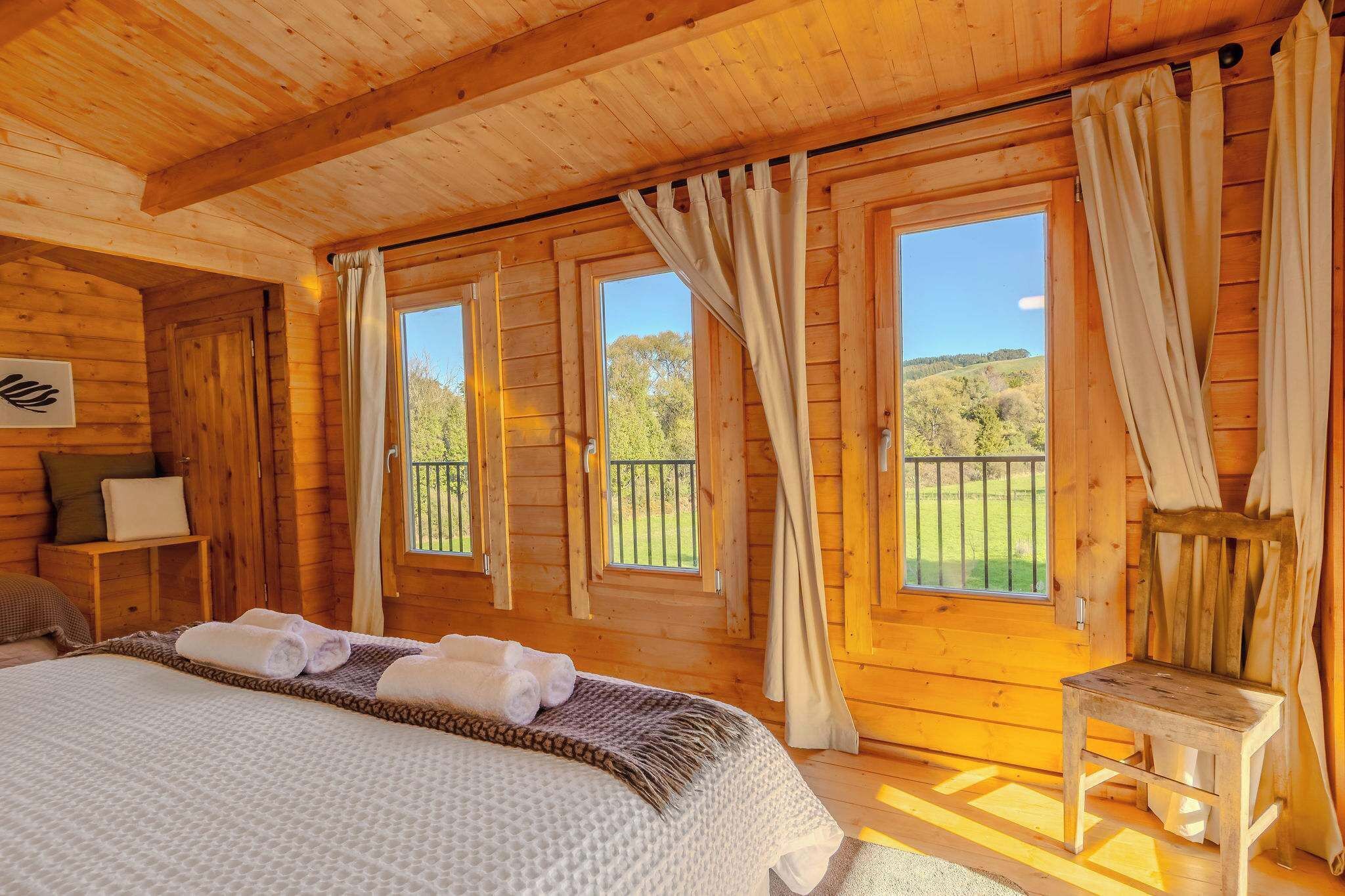 Una habitación de cabaña de madera con una cama, dos ventanas con cortinas, una silla y una mesita.