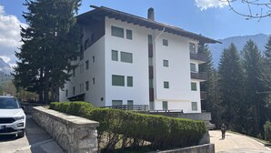 Un edificio de varios pisos con fachada blanca y contraventanas verdes, rodeado de árboles y montañas.
