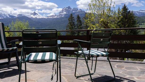 Dos sillas verdes con cojines a rayas sobre un patio de piedra con vistas a un paisaje montañoso con picos nevados y un cielo azul despejado.