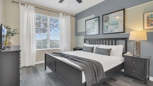 Une chambre moderne avec un grand lit, des tables de chevet, une petite table, un ventilateur de plafond et une fenêtre avec des stores.