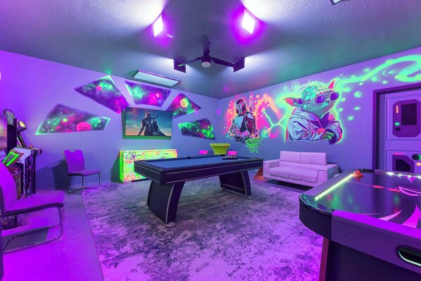 Une pièce avec une table de billard, des œuvres d’art murales néon, un canapé blanc et un ventilateur de plafond.