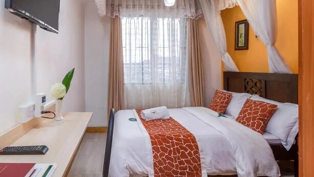 A hotel room with a bed, a desk with a remote control, a vase with flowers, and a window with curtains.