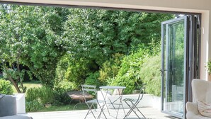 A modern patio with a glass door leading to a lush garden. There is a dining table with chairs and a sofa. - Hathaway Lane (Stratford-upon-Avon)