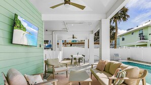 A poolside area with a sofa, chairs, and a table. There is a TV mounted on the wall displaying a beach scene.