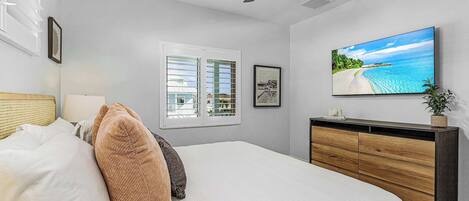 A bedroom with a bed, a ceiling fan, a TV, a window with blinds, a bedside table, and a dresser.