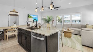 A modern kitchen with a central island, a dining area with a wooden table and chairs, a sofa, and a large window with blinds.