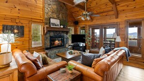 A cozy cabin interior with a stone fireplace, leather furniture, and a television. - Heinrich's Hideaway Blue Ridge, GA (MORGANTON)