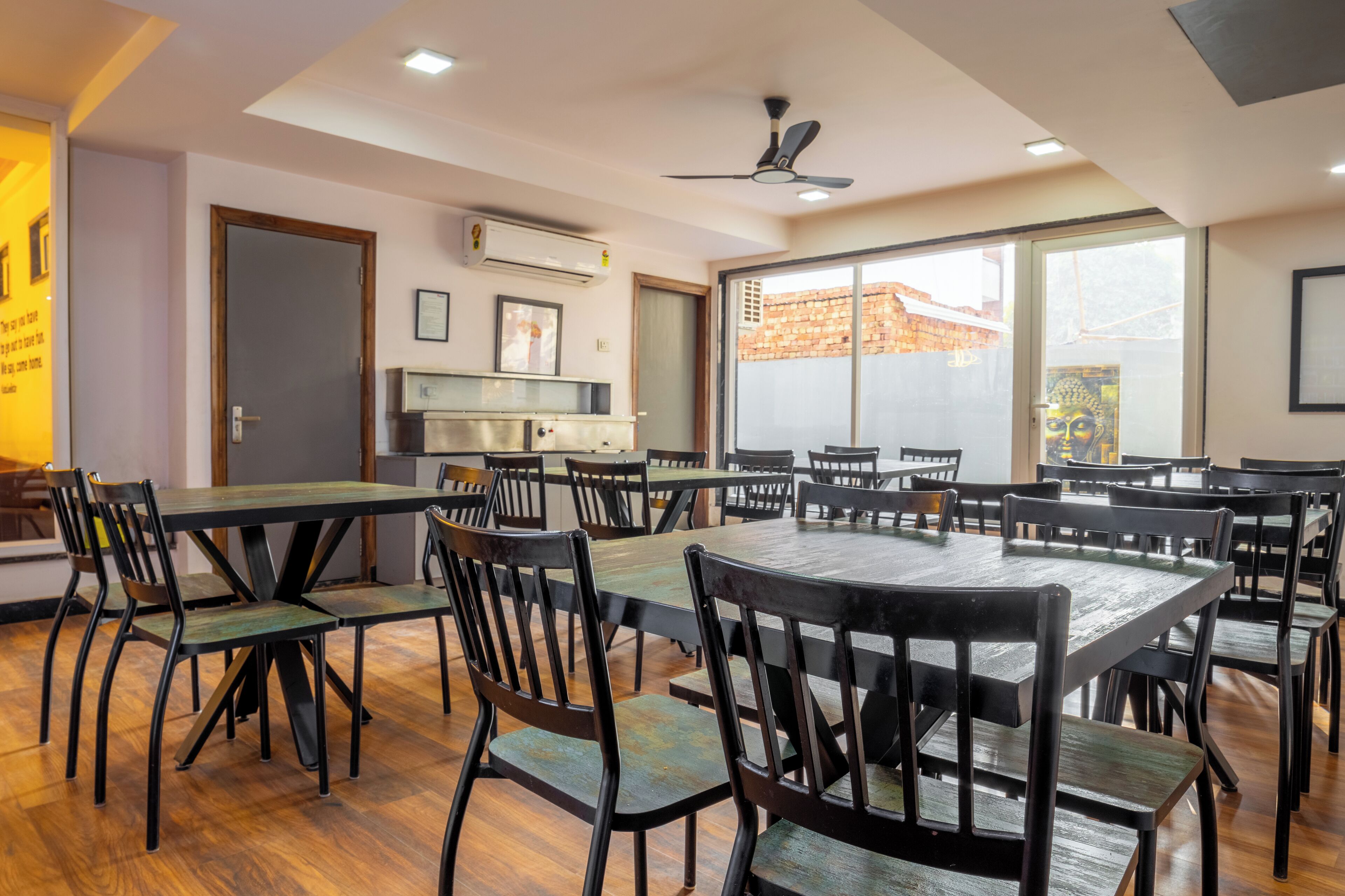 Un vaste espace dédié à la salle à manger, avec plusieurs tables et chaises, un ventilateur de plafond et de grandes fenêtres offrant une vue sur l’extérieur.