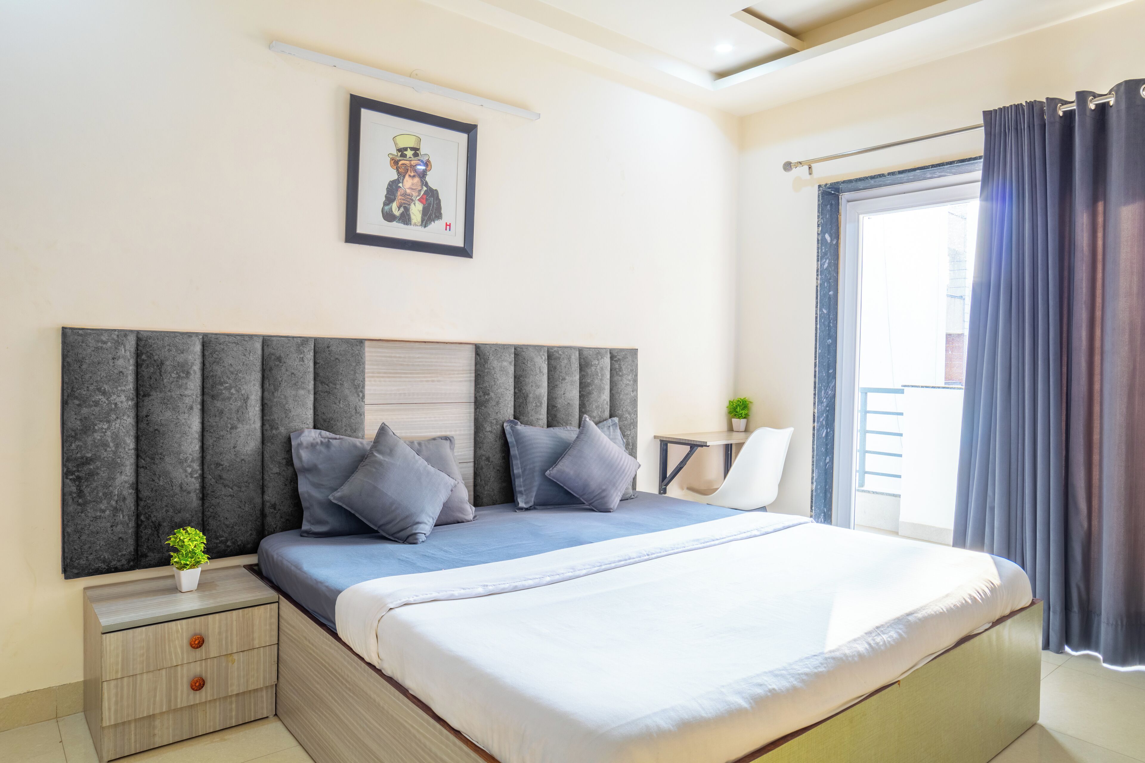 A bedroom with a large bed, a nightstand with drawers, a framed picture on the wall, and a window with curtains.