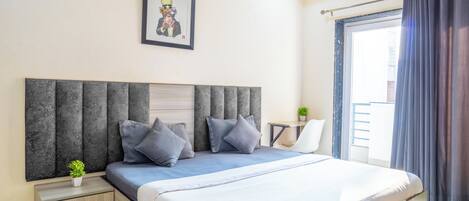 A bedroom with a large bed, a nightstand with drawers, a framed picture on the wall, and a window with curtains.