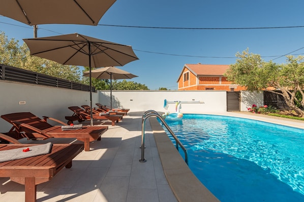 A poolside area with wooden loungers and umbrellas, a swimming pool, and a building in the background.