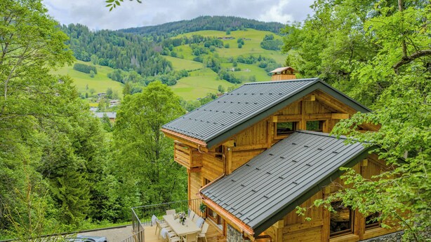 A wooden cabin with a hot tub and a dining area, surrounded by lush greenery and a view of rolling hills.