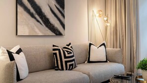 Un salon moderne avec un canapé gris, des coussins aux motifs noir et blanc, une grande peinture abstraite, un grand lampadaire et une table basse en verre avec une télécommande.