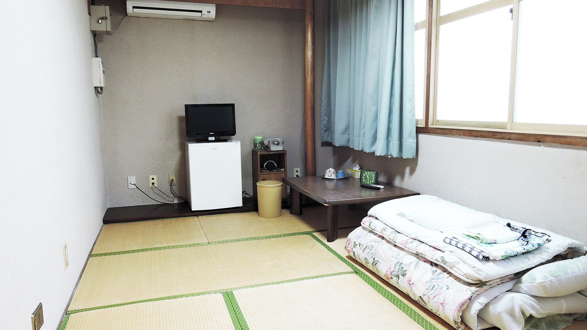 Une petite pièce avec un sol recouvert de tatamis, un lit avec une literie à motifs floraux, un bureau avec une télévision, un petit radiateur et une fenêtre avec un rideau.