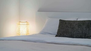 Un letto rifatto con lenzuola bianche e un cuscino scuro, una lampada cilindrica con decorazioni intrecciate che illumina la parete, e un soffitto bianco liscio.