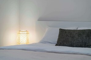 A neatly made bed with white linens and a single dark-colored pillow, a cylindrical lamp with a woven design casting light on the wall, and a plain white ceiling.
