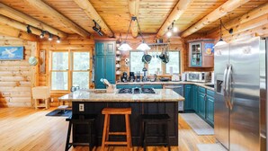 A log cabin kitchen with wooden beams, a central island with stools, a refrigerator, and a window overlooking greenery. - Wingspan Cabin (Keedysville)