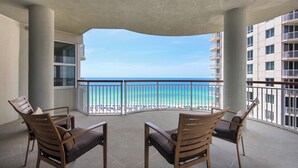 A balcony with two wooden chairs and a table, overlooking a beach and a hotel building. - Beach Colony West 10E 2BR Gulf Front 2/Beach Service & Onsite Management (Navarre)