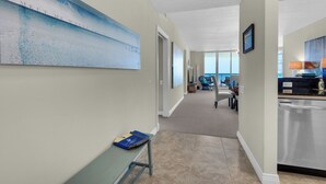 A hotel hallway with a large abstract painting on the left, a bench with a book, and a view into a dining area with chairs and a table. - Beach Colony West 10E 2BR Gulf Front 2/Beach Service & Onsite Management (Navarre)