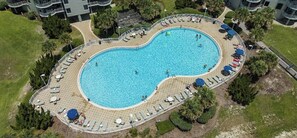 Vista aérea de una piscina con forma de corazón rodeada de sillones de descanso y sombrillas, con zonas ajardinadas y edificios al fondo.