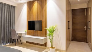 A modern living room with a flat-screen TV mounted on a wooden panel wall, a white console table, a grey sofa, a potted plant, and a wooden door. - Housr Sector 56 Gurgaon Sushant Lok 2 (Gurugram)