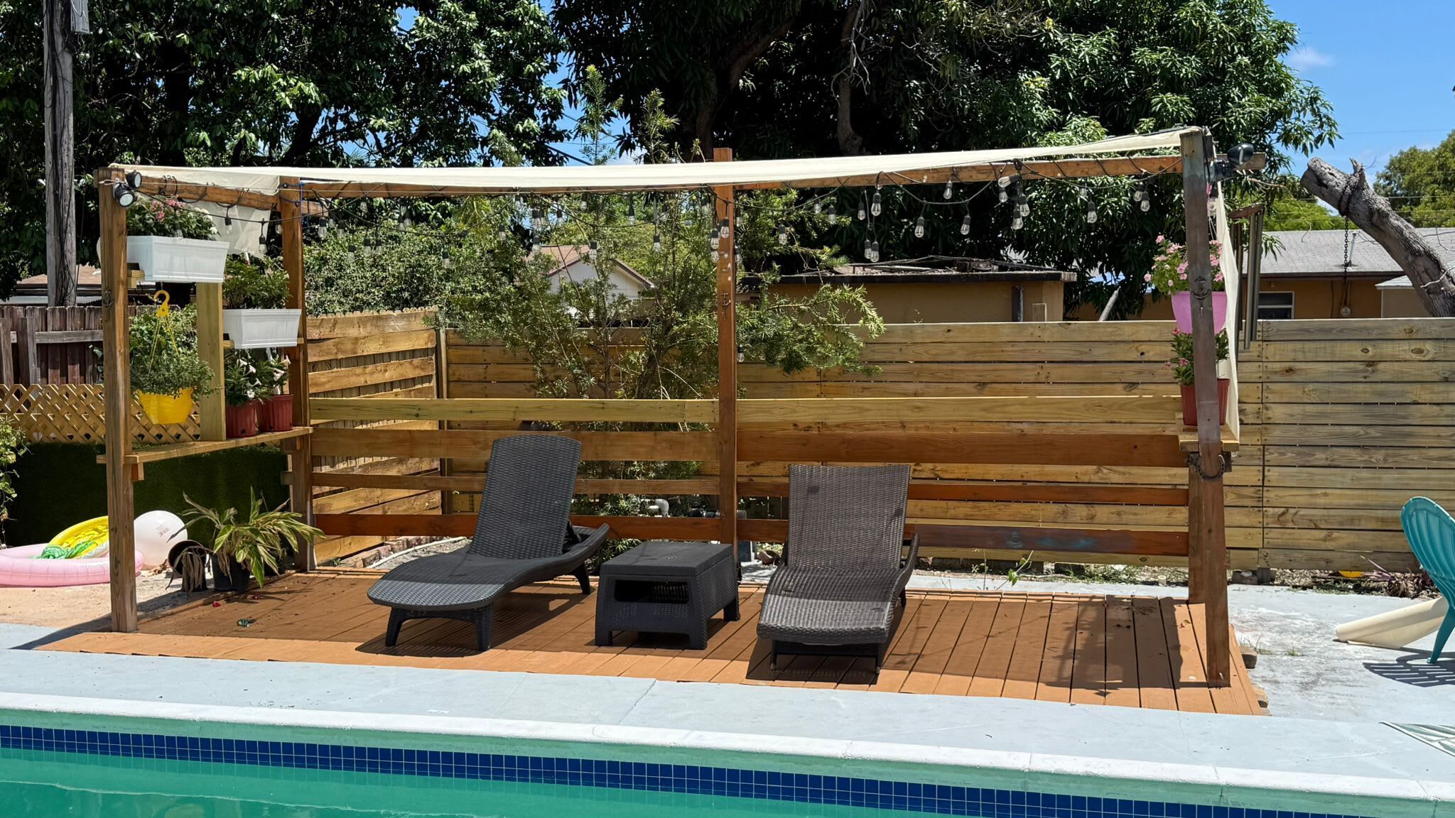 Zona junto a la piscina con terraza de madera, dos sillas reclinables, una mesita y una pérgola con luces colgantes.