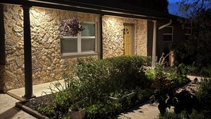 Une maison en pierre avec une porte jaune, une fenêtre agrémentée d’une jardinière et un jardin où poussent différentes plantes.