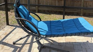 Une chaise longue avec un coussin bleu et un auvent, placée sur une terrasse en briques, avec une clôture en bois en arrière-plan.