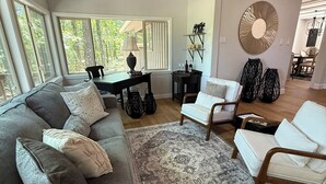 Ein geräumiges Wohnzimmer mit einem großen grauen Ecksofa, einem weißen Sessel und einem gemusterten Teppich. Im Hintergrund ist ein Essbereich mit einer Chandelier und einem wandmontierten Spiegel zu sehen.